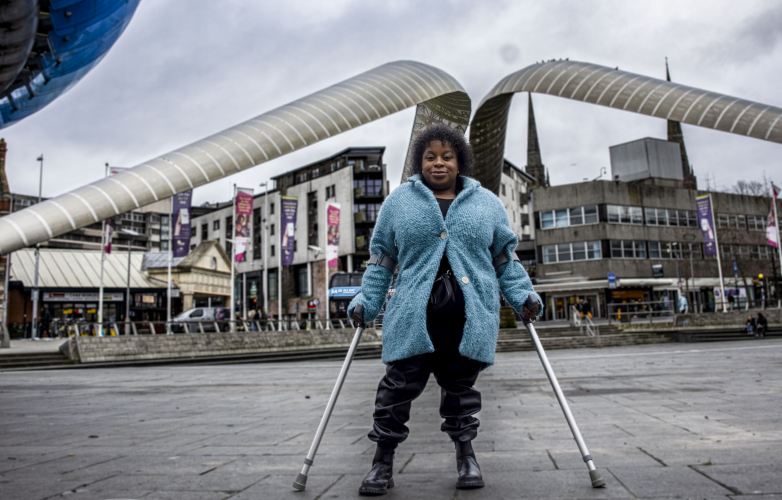 A woman stands holding crutches with buildings behind her and a grey cloudy sky She is wearing a bright blue warm jacket and is smiling facing the camera