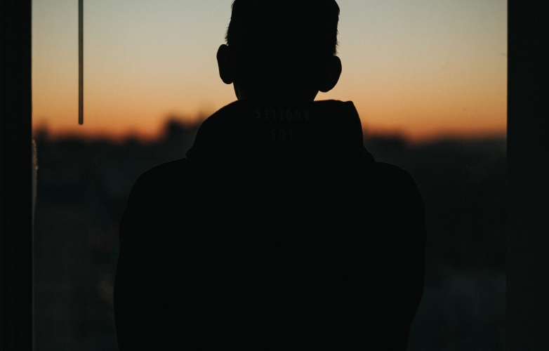 A man stands at a window at dusk his back is turned and we just see his outline