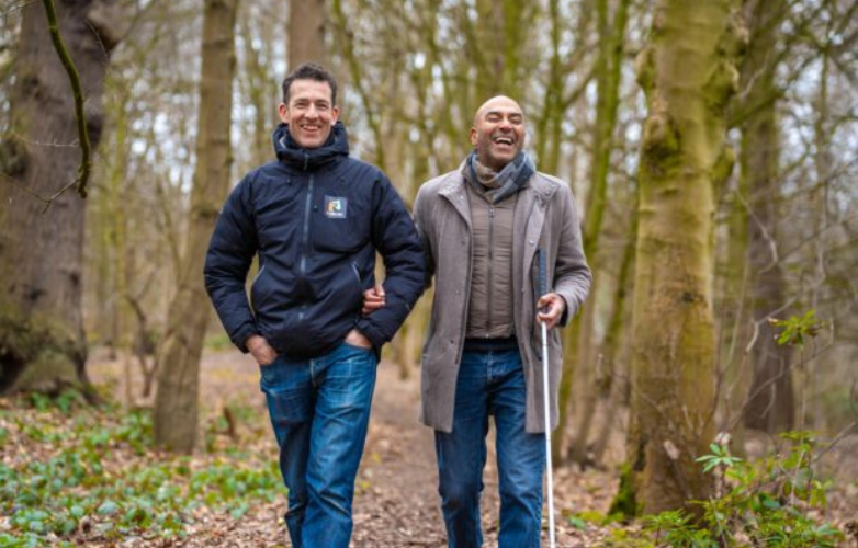 Two men outside walking in the woods One at the front is guiding the other who uses a white cane They are both laughing together