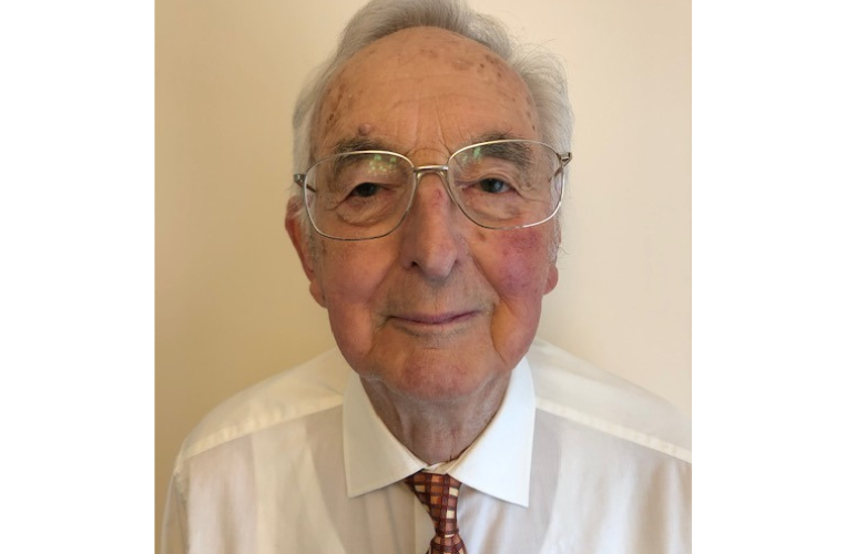 Close up of Arthurs face and shoulder looking directly into the camera with a soft smile and large wire rimmed glasses he wears a white shirt and red checked tie
