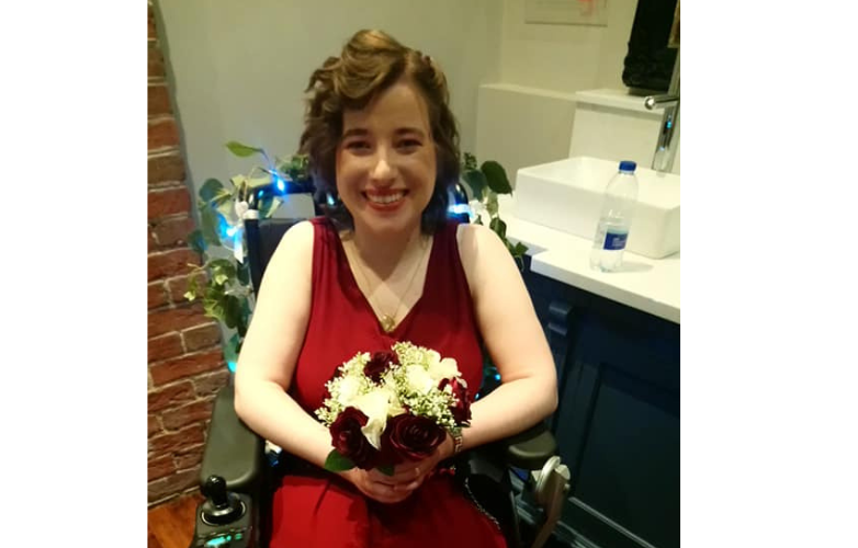 Becca wears a silky red dress and holds a boquet of flowers sitting down smiling 