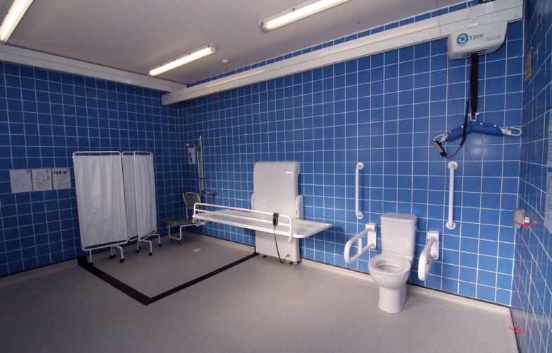Image shows the inside of a Changing Places toilet. It is a large space with blue tiled walls and equipment such as curtains, a hoist, changing bench and toilet 