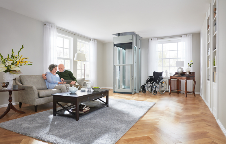 Picture of a couple sitting on a sofa in a living room with a homelift in the corner. There is also a wheelchair beside the homelift