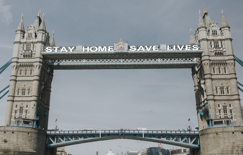 Image shows a full view of tower bridge in London, it has a banner across it which reads 'stay home save lives'