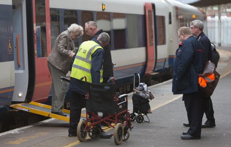 A picture of an older woman being assisted off a train via a ramp. There are four men assisting her. 