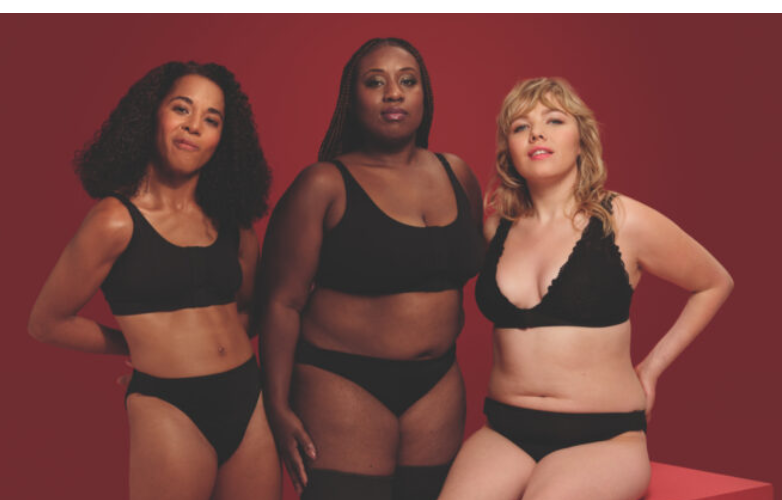 Three young woman stand against a deep red background they all wear plain black underwear and stand looking into the camera