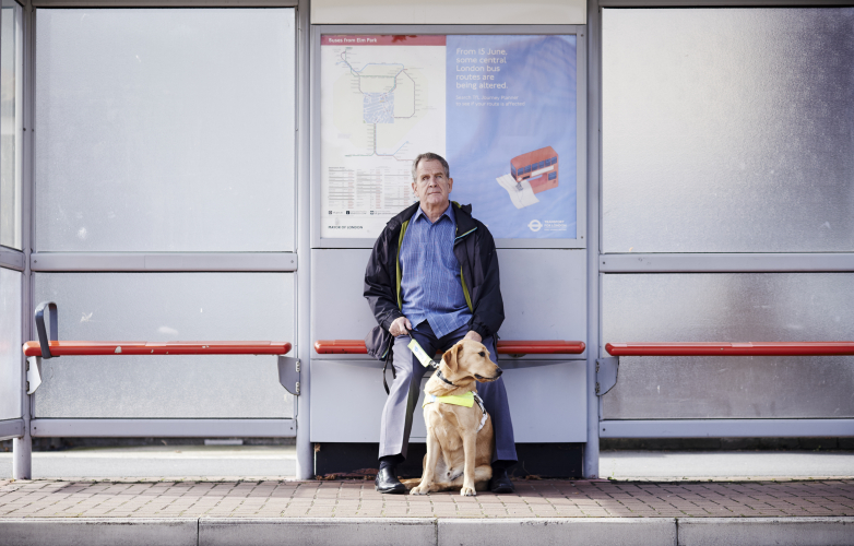 A visually person waiting at a bus stop with his guide dog