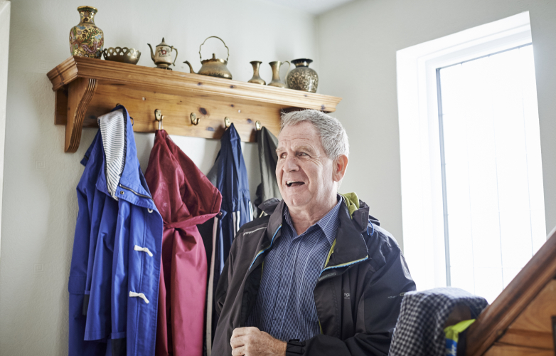 Image shows a visually impaired man and member of RiDC's panel wearing his coat at home, by the front door. He is smiling. 