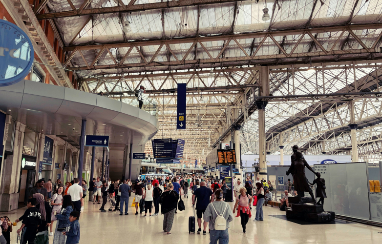 Busy indoor station scene with lots of people walking and standing in the foreground Station signs are in the distance and a noticebaord with train times Above iron cladding covers the ceilings 