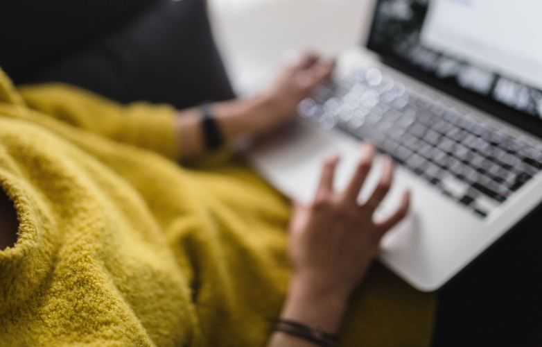 Image shows a woman sitting on sofa at home using a computer 