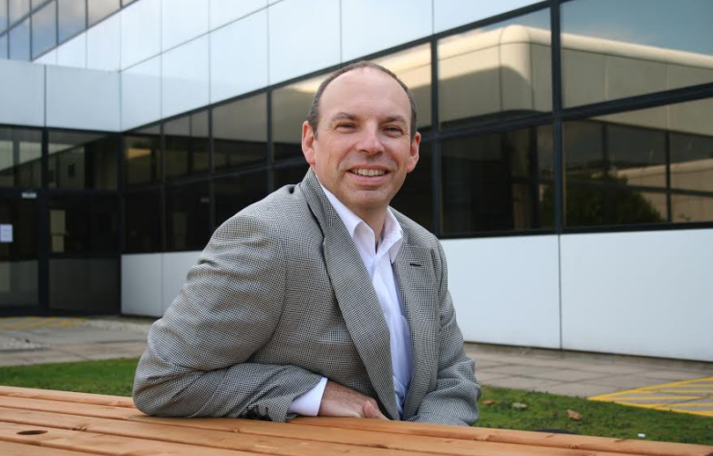 A man sitting outside in front of a modern building he wears a suit and smiles at the camera